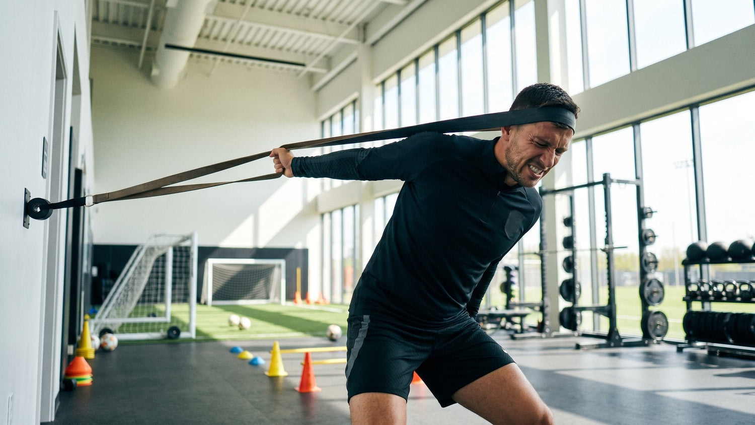Male soccer player performing resistance band neck training exercises in a professional sports facility