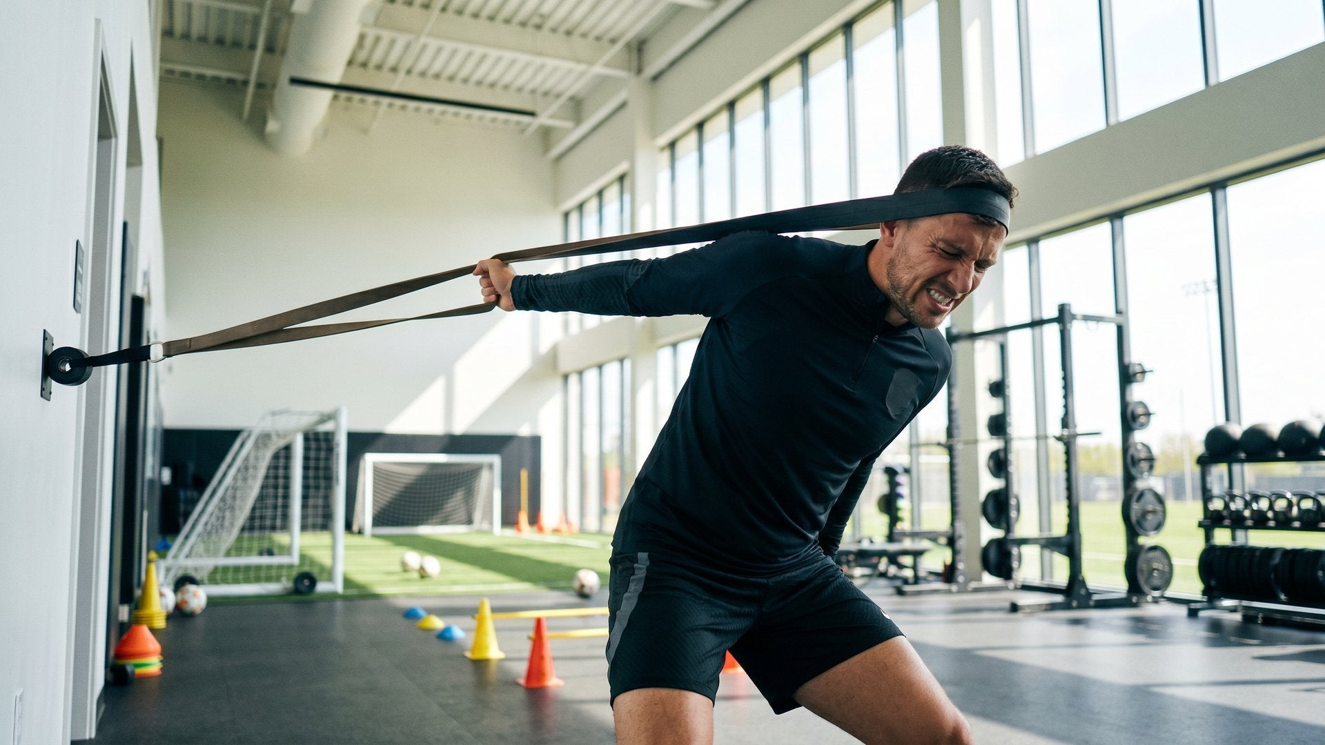 Male soccer player performing resistance band neck training exercises in a professional sports facility